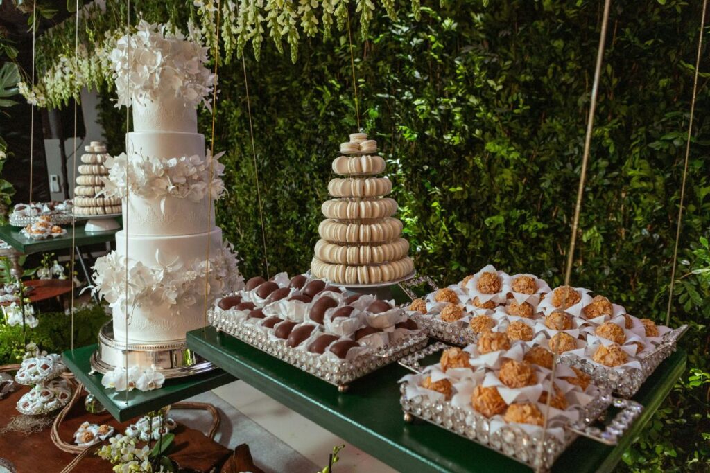 Luxurious wedding dessert table with multi-tiered white floral cake, macaron tower, trays of golden pastries and chocolate treats, styled with crystal-edged trays and lush greenery backdrop.