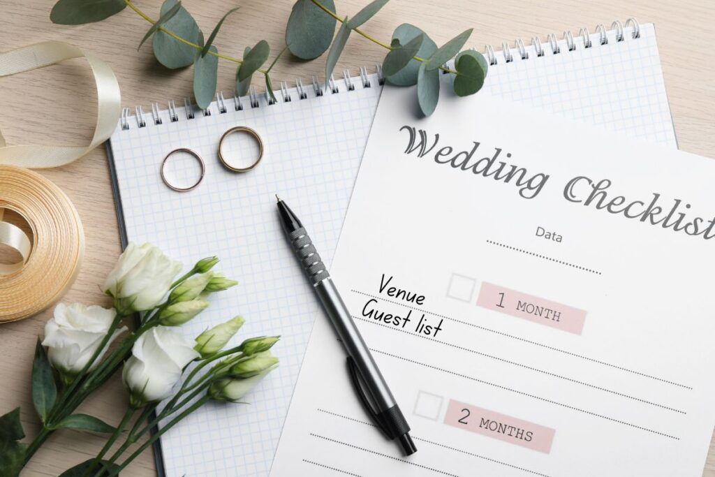 Flat lay of a wedding planning checklist with notebooks, pen, wedding rings, ribbon, and flowers on a wooden table, symbolizing organized stress‑free wedding preparation.