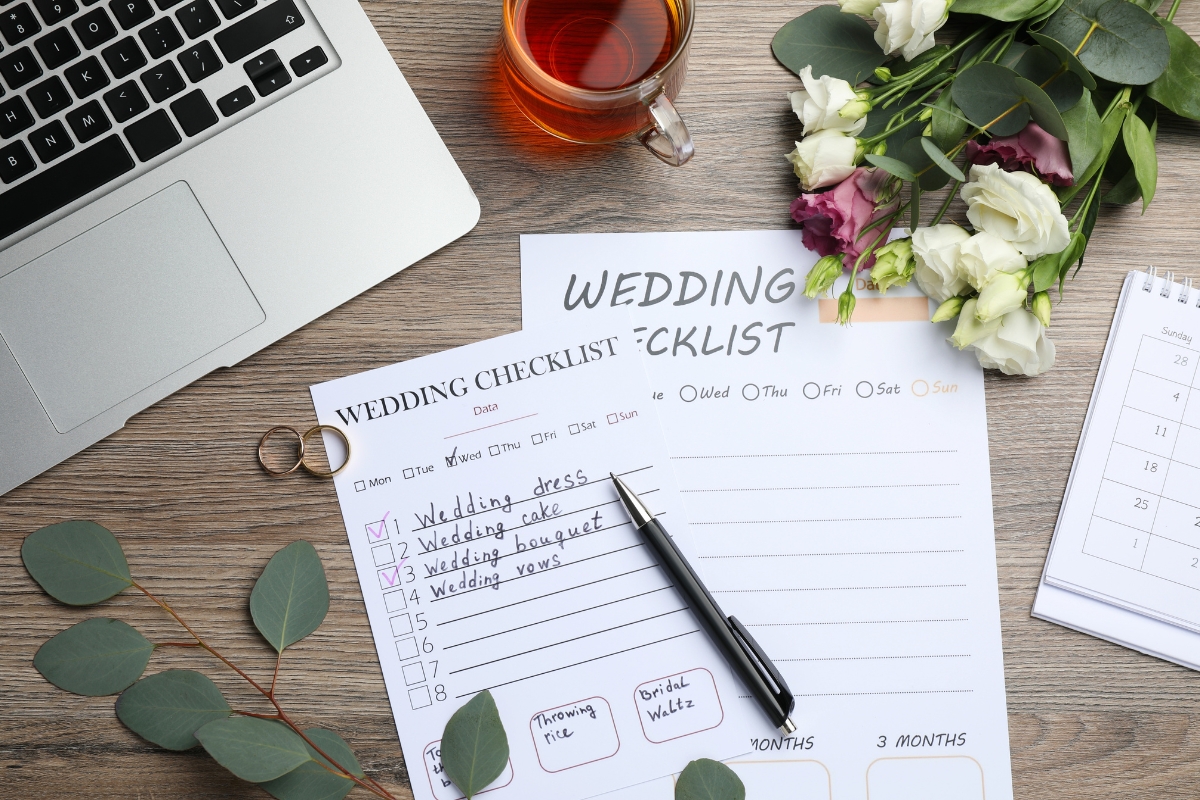 Wedding planning timeline workspace with laptop, tea, bouquet, rings, and calendar on wooden table, centered around wedding checklist sheets with handwritten tasks and pen, symbolizing organized preparation for the big day.